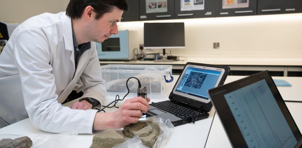 A heritage scientist in a white coat in a lab, scanning a piece of stone in front of a laptop
